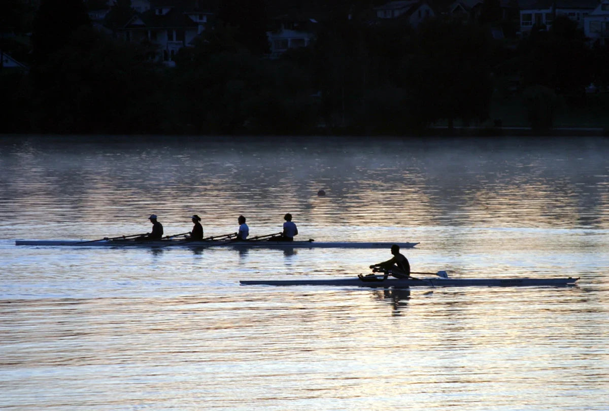 Silhouetted rowers on calm water at dusk, with two racing boats visible against a dark shoreline and golden reflections on the lake.