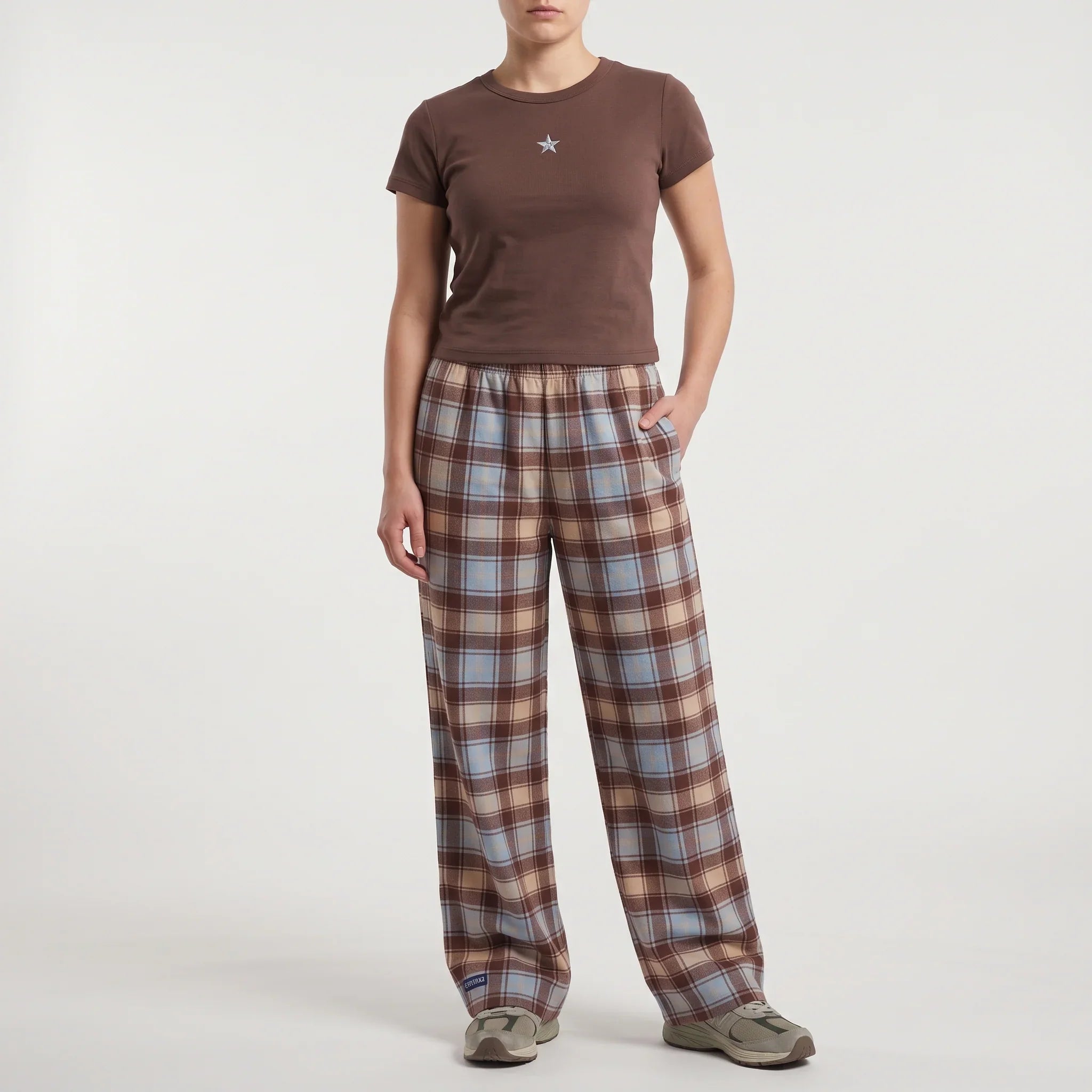 A model wearing a brown short-sleeve t-shirt with a small embroidered star on the chest and brown plaid lounge pants with light blue and beige stripes, standing against a light gray studio background.