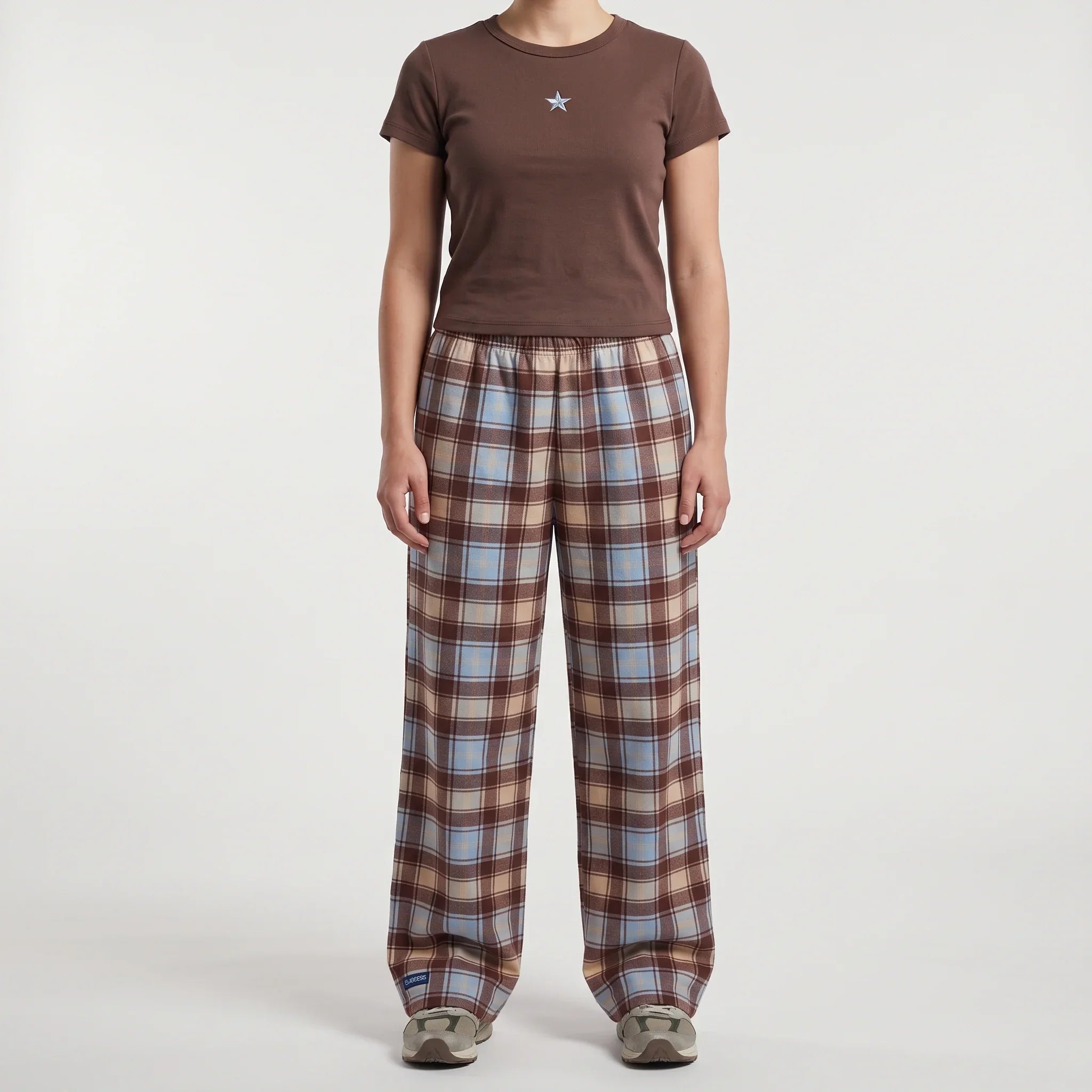 A model wearing a brown short-sleeve t-shirt with a small star logo on the chest and brown plaid lounge pants with blue and beige stripes, standing against a light gray background.