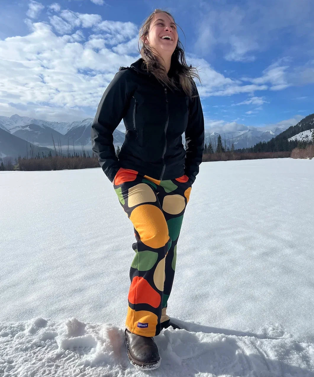 Person wearing colorful pants and a black jacket standing on a snow-covered landscape with mountains in the background.