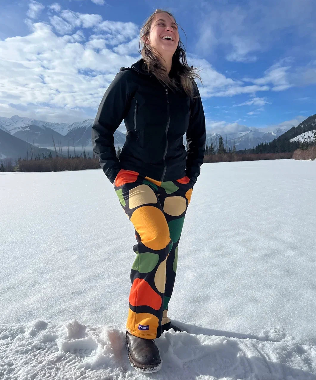 Person wearing colorful pants and a black jacket standing on a snow-covered landscape with mountains in the background.