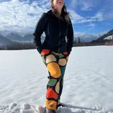 Person wearing colorful pants and a black jacket standing on a snow-covered landscape with mountains in the background.